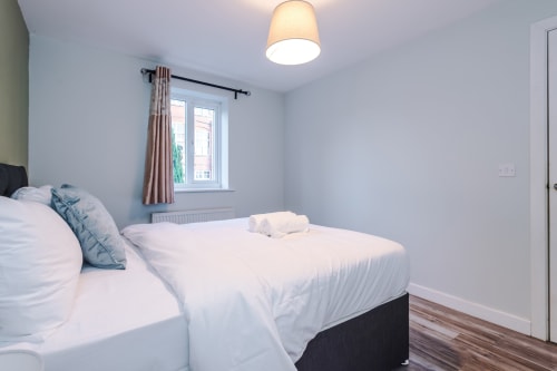 Modern bedroom at Vidale Court villa in Manchester with double bed, blue pillows, window with curtains, ceiling light, and