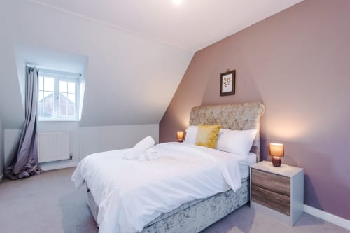 Cozy attic bedroom at Vidale Court Manchester villa with mauve walls, white bedding, yellow accent pillow, and bedside lamps