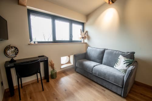 Studio moderne à Lieu-dit Boulage avec canapé gris, bureau, horloge, grandes fenêtres et parquet en bois à Marquefave.
