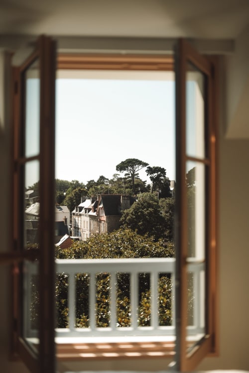 Portes en bois ouvertes révélant la vue sur Dinard avec bâtiments historiques et arbres depuis le balcon de la chambre de l'Hôtel Saint Enogat