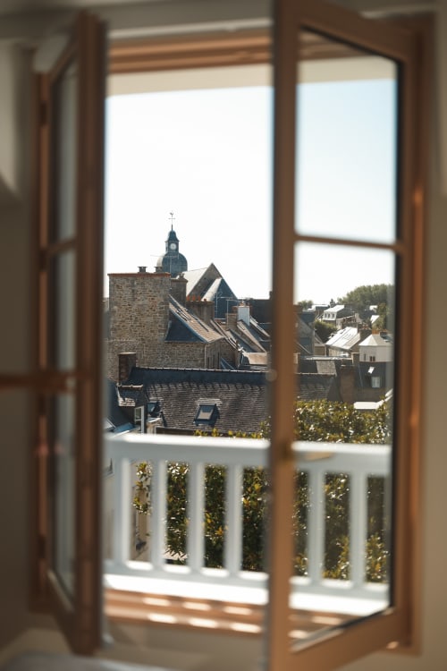 Fenêtre en bois ouverte sur la ville de Dinard avec clocher d'église et toits historiques depuis une chambre de l'Hôtel Saint Enogat