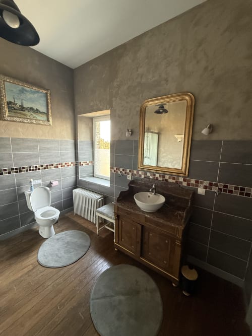Salle de bains spacieuse du Château De Montbrault avec vanité vintage, toilettes blanches, murs en carrelage foncé et miroir doré