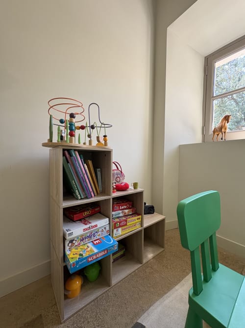 Salle de jeux pour enfants avec étagère en bois rangant livres et jeux, jouet labyrinthe de perles, et chaise verte à la Conciergerie Maurin
