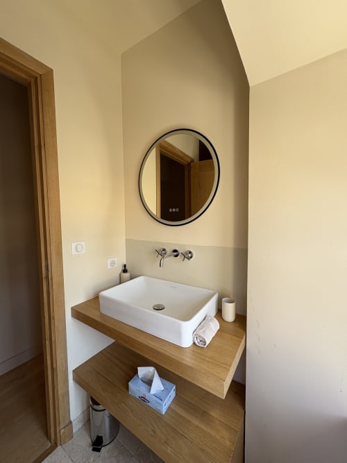 Salle de bain moderne avec lavabo blanc, miroir rond noir, meuble en bois et robinet mural à la Conciergerie Maurin