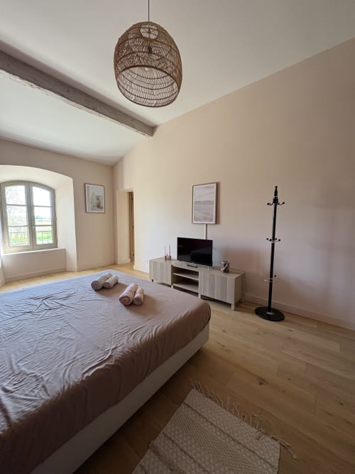 Chambre spacieuse à la Conciergerie Maurin à Arles avec lit, luminaire suspendu tissé, TV, bureau et parquet.
