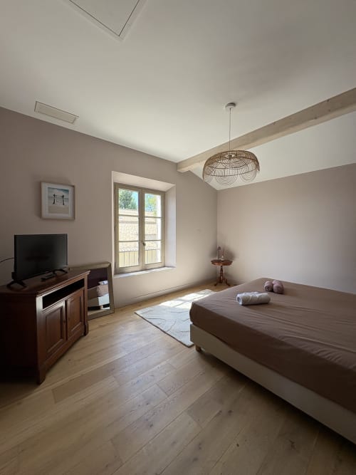 Chambre spacieuse à la Conciergerie Maurin à Arles avec lit, TV, bureau en bois, portes vitrées donnant sur balcon et suspension en osier