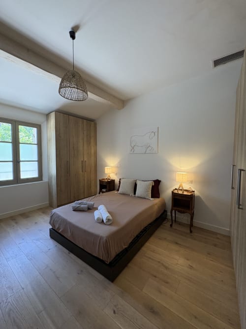 Chambre moderne à la Conciergerie Maurin à Arles avec armoire en bois, lit aux serviettes roulées, suspension luminaire et vue sur le jardin