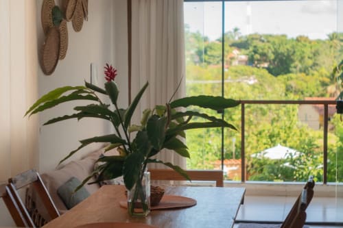 Área de refeições da Vila Pitangueira com planta em vaso, mesa de madeira e vistas panorâmicas da paisagem de Uruçuca através de grandes janelas