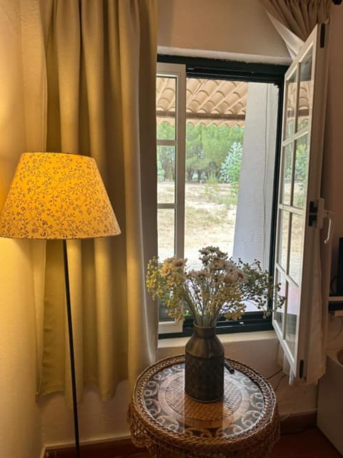 Quarto aconchegante na Monte da Tojeirinha com luminária amarela, mesa redonda, flores secas e portas francesas abertas com vista para os jardins