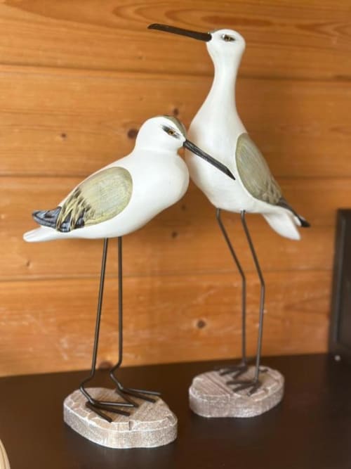 Duas esculturas decorativas de pássaros branco e verde sálvia com pernas longas e finas em bases de madeira, expostas no interior de Monte da