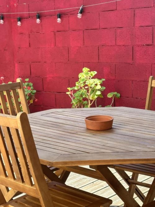 Mesa de jantar em madeira com cadeiras no terraço da quinta Monte da Tojeirinha, parede de tijolos vermelhos vibrantes, plantas em vasos