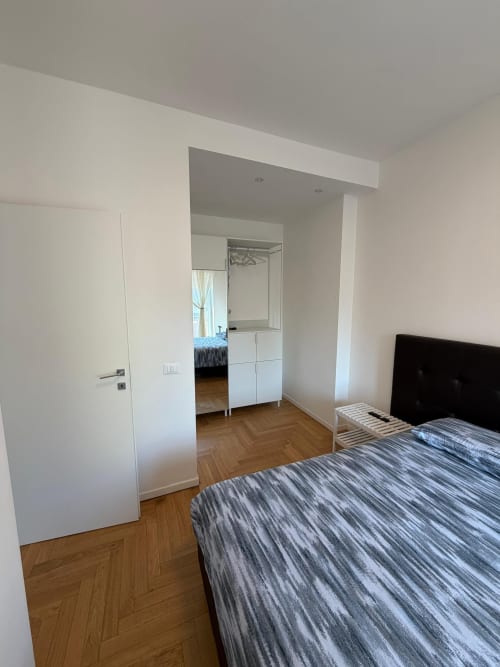 Camera da letto moderna in appartamento milanese con letto a righe, porta bianca, pavimento in legno e accesso al bagno ensuite