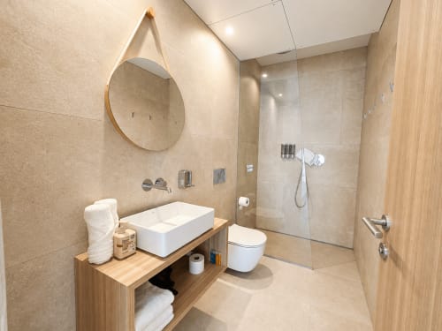 Baño moderno en Holibai ModulSuites de Baiona con lavamanos de cerámica, espejo redondo, ducha a ras de suelo y tocador de madera