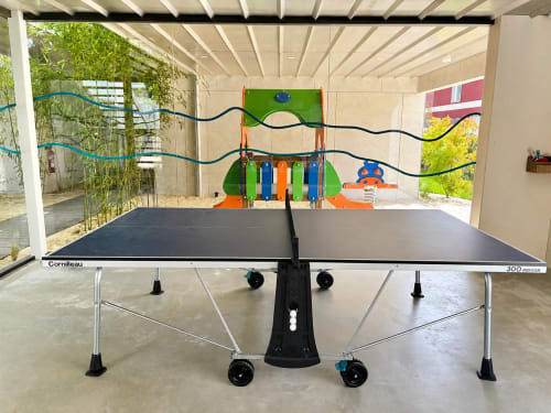 Mesa de tenis de mesa en zona cubierta de recreo en apartamentos Holibai, Baiona con coloridos equipos de juego visibles a través del cristal
