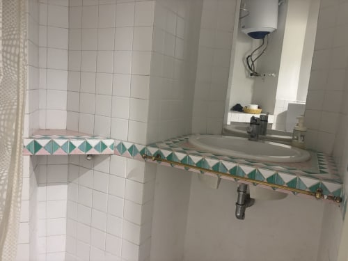Bagno con pareti piastrellate bianche, bordo decorativo verde acqua e rosa, lavabo, specchio e scaldabagno a muro