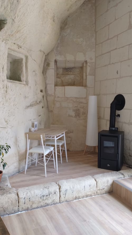 Chambre La Cave 1872 aux Troglos de Beaulieu : murs en pierre brute, chaises blanches, poêle en bois noir et mobilier rustique