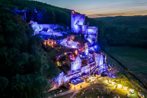 Ruines du château médiéval illuminées au crépuscule à Carsac-Aillac, France, avec collines boisées et visiteurs explorant les lieux