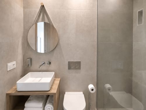 Cuarto de baño moderno en apartamento Holibai, Baiona con lavabo tipo vessel blanco, espejo redondo, ducha, inodoro y azulejos beige