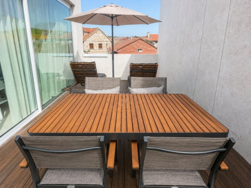 Terraza moderna con suelo de madera, mesa de comedor, tumbonas, sombrilla y vistas de la ciudad de Baiona en el apartamento hotel Holibai