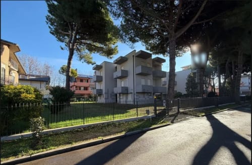 Moderno edificio residenziale a Cervia con giardino verde, area recintata e alti pini sotto cielo sereno