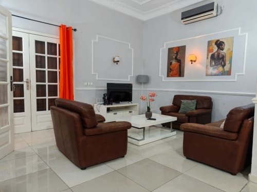 Salon moderne à La Villa des Pins à Libreville avec canapé en cuir marron, murs blancs, téléviseur et tableaux encadrés