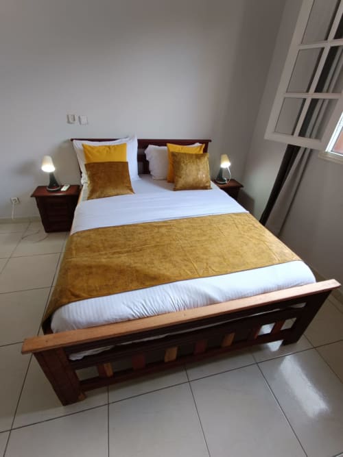 Chambre élégante à La Villa des Pins, Libreville avec accents dorés, lit en bois, draps blancs et vue sur le jardin