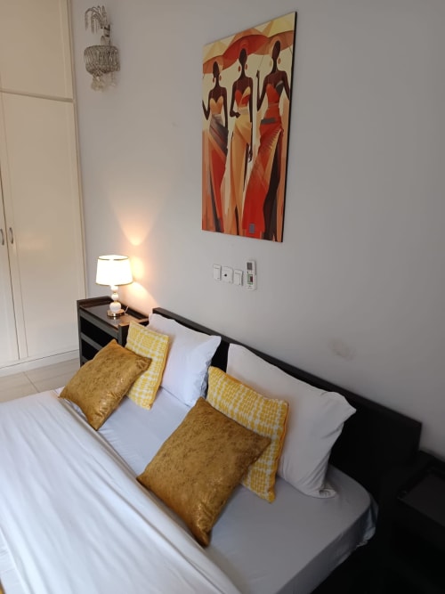 Chambre à la Toscane à La Villa des Pins avec coussins dorés, lit à cadre noir, lampe de chevet et tableau coloré sur mur blanc