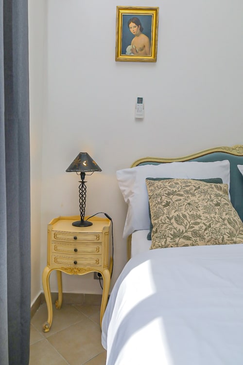 Chambre élégante à La Villa des Pins avec table de nuit dorée, lampe noire, coussin à motifs et portrait encadré sur mur blanc