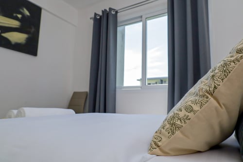 Chambre moderne à La Villa des Pins à Libreville avec lit blanc, coussins décératifs, rideaux gris et vue sur le jardin