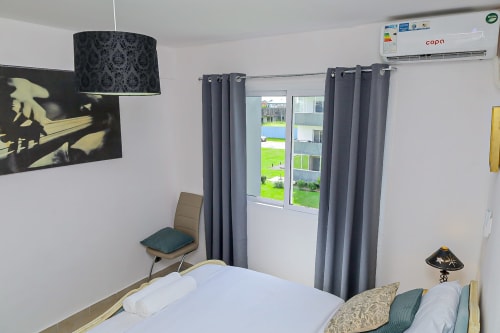 Chambre moderne à La Villa des Pins à Libreville avec vue jardin, lit blanc, rideaux gris, climatisation et décoration contemporaine