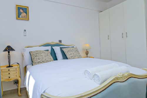 Chambre élégante à La Villa des Pins avec lit doré orné, draps blancs, coussins à motifs et armoires blanches modernes