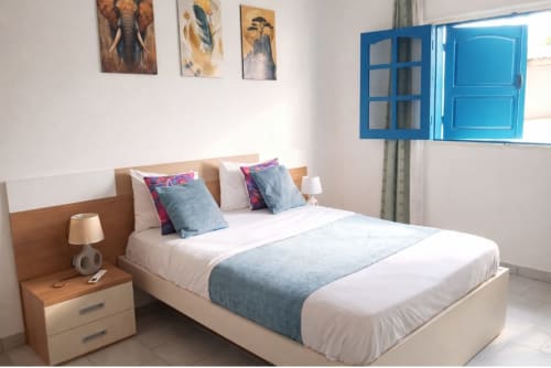 Chambre moderne et lumineuse à La Villa des Pins à Akanda avec lit queen, table de nuit en bois, fenêtre aux volets bleus