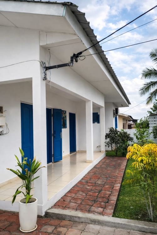 Villa blanche aux portes bleues et terrasse couverte à La Villa des Pins à Akanda, avec allée en briques et plantes tropicales