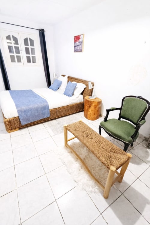 Chambre à La Villa des Pins à Akanda avec lit en rotin, linge bleu, fauteuil vert et banc tressé