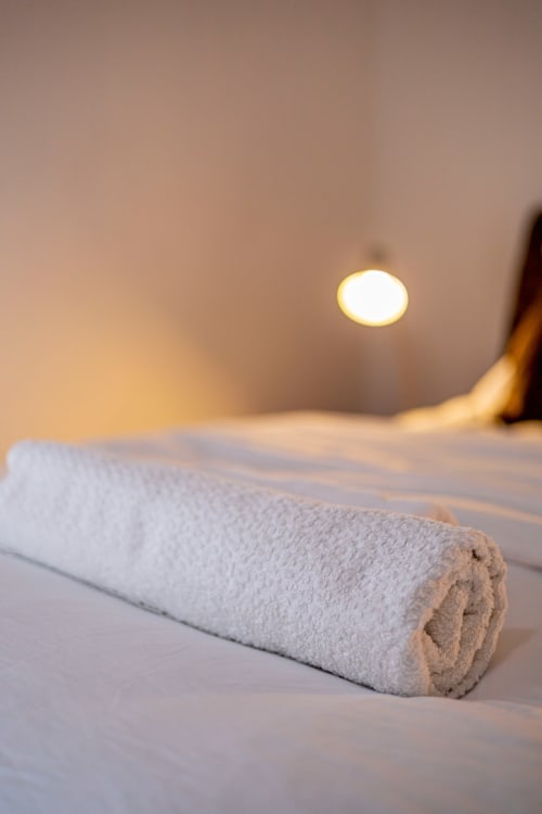 Serviette blanche roulée sur le lit, éclairée par la lumière douce du plafonnier à l'appartement La Villa des Pins