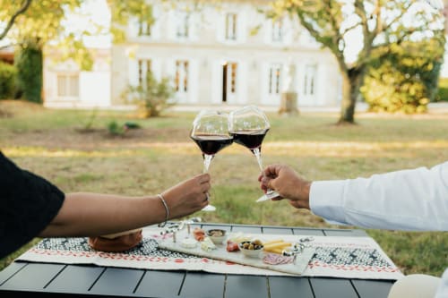 Dos copas de vino brindando en el Château Alto Cormeil en Saint-Émilion, con embutidos y una casa señorial blanca de fondo