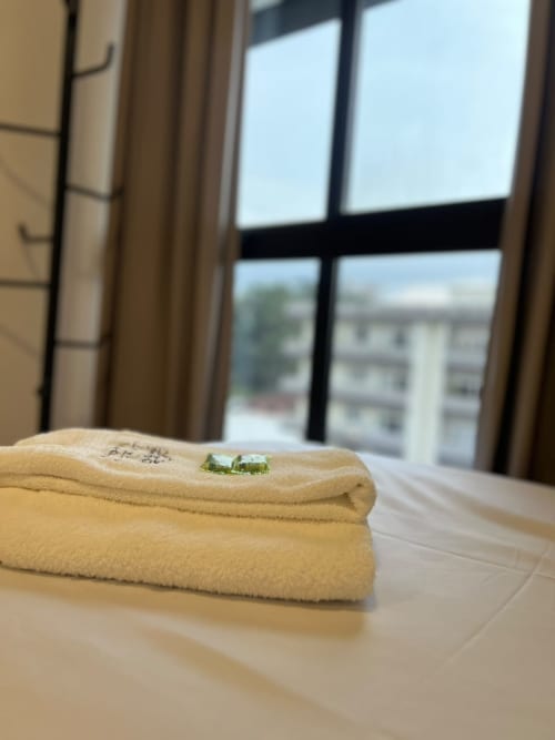Toalhas dobradas com pequenas plantas verdes em mesa de madeira, janelas com vista da cidade e cortinas no apartamento Bora Bora Hospedagens em