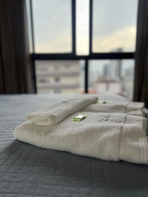 Toalhas brancas e fofas com detalhe de folha verde na cama, vista da cidade pela janela no apartamento BORA BORA HOSPEDAGENS em Joinville