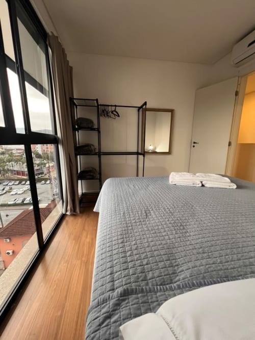 Quarto moderno em apartamento em Joinville com vista da cidade, cama queen, piso de madeira, prateleiras pretas e grande janela com vista