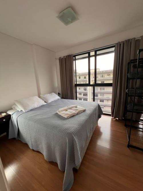 Quarto moderno em apartamento duplex em Joinville com vista da cidade, cama, piso de madeira e varanda com vista para o shopping Mueller