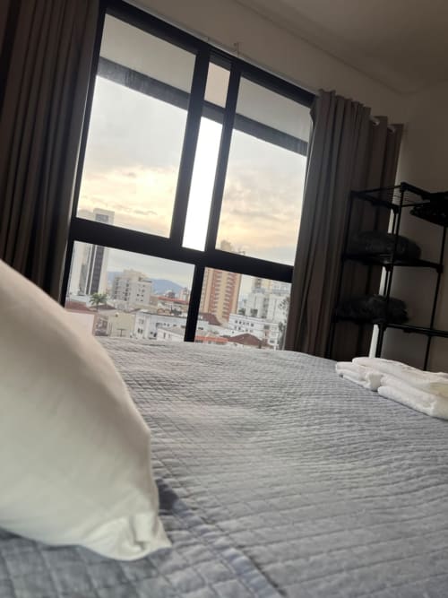 Quarto moderno na BORA BORA HOSPEDAGENS, Joinville, com vista da cidade através de grandes janelas e toalhas dobradas na cama