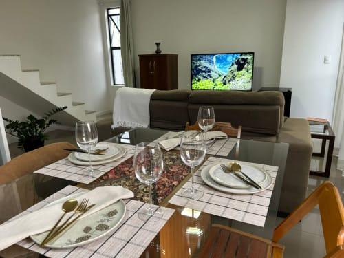 Área de refeições em apartamento de Joinville com mesa posta, sofá cinza, TV e escada ao fundo
