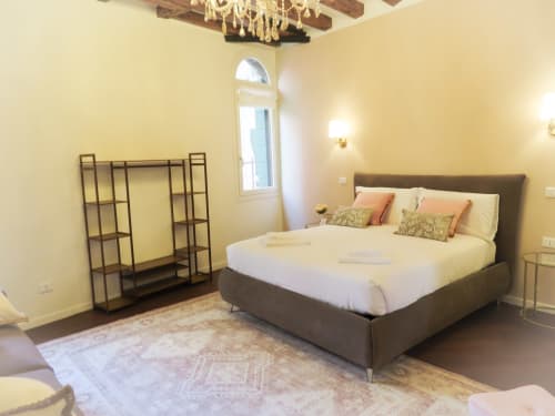 Luxury suite bedroom in Verona with grey upholstered bed, yellow walls, wooden beams, arched window, and decorative shelving
