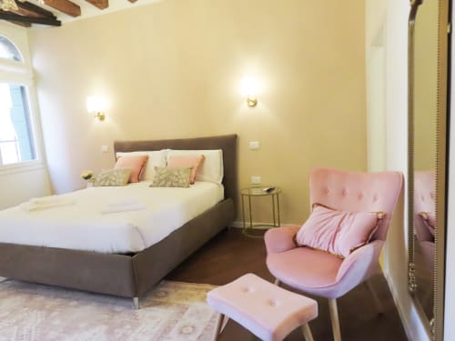 Luxury suite bedroom in Verona with gray bed, pink armchair, yellow walls, wooden beams, and river view windows.