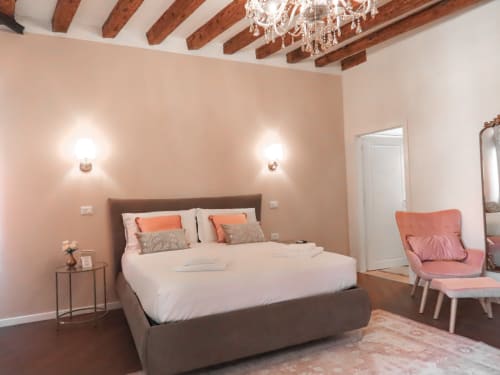 Luxury suite bedroom in Verona with exposed wooden beams, pink walls, dark bed, crystal chandelier, and river view access