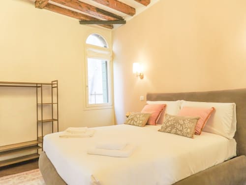 Bright bedroom at San Marco Luxury Suites in Verona with queen bed, decorative pillows, arched window, wooden beams, and wall