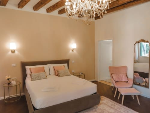 Luxury bedroom suite in Verona with elegant furnishings, crystal chandelier, white bedding, and pink accent chair overlooking