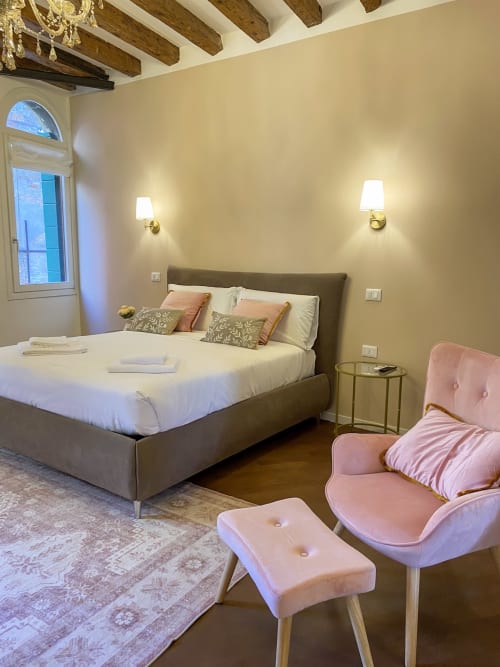Elegant bedroom suite in Verona with grey upholstered bed, pink velvet seating, exposed wooden beams, and river view window