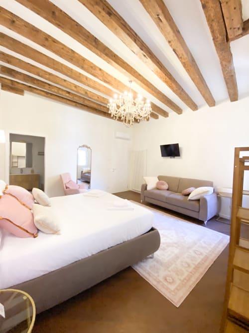 Luxury suite in Verona with exposed wooden beams, white walls, queen bed with pink pillows, sofa, crystal chandelier, and