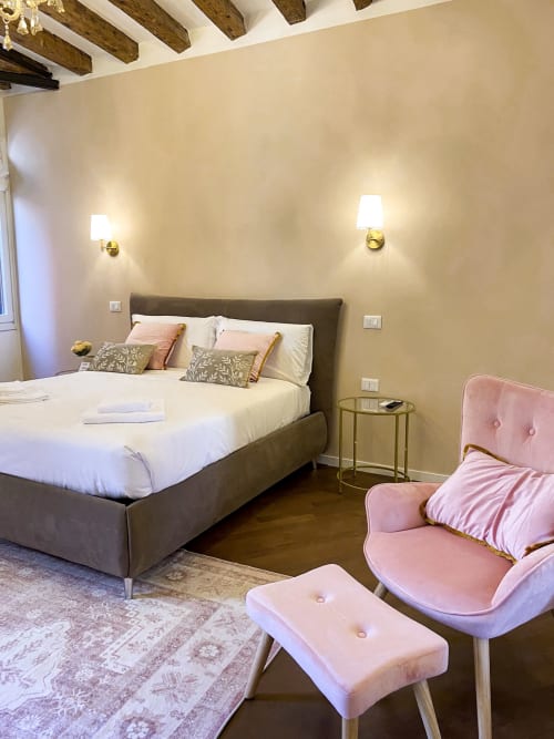 Elegant bedroom in San Marco Luxury Suite, Verona with gray upholstered bed, pink velvet chair, exposed wooden beams, and